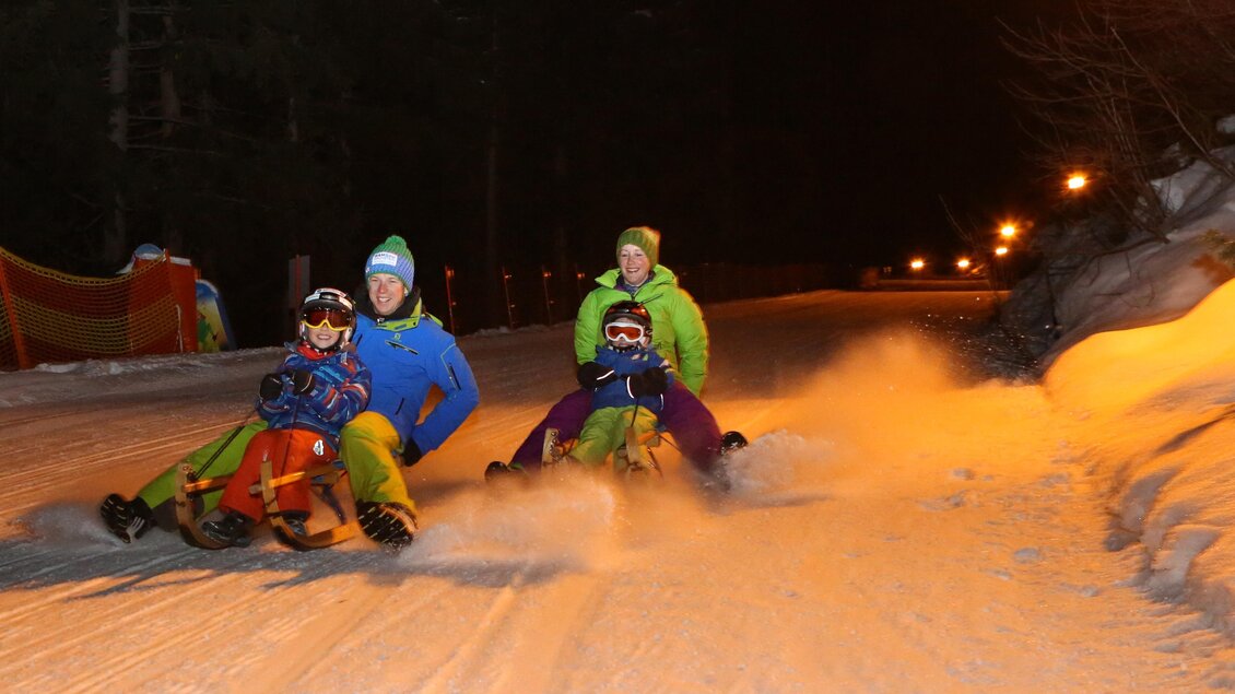 Eine Gruppe von vier Personen fährt nachts mit Schlitten auf einer schneebedeckten Piste. Umgeben von Dunkelheit sind Lichter im Hintergrund sichtbar, die eine fröhliche Stimmung verbreiten. | © Hans-Peter Steiner