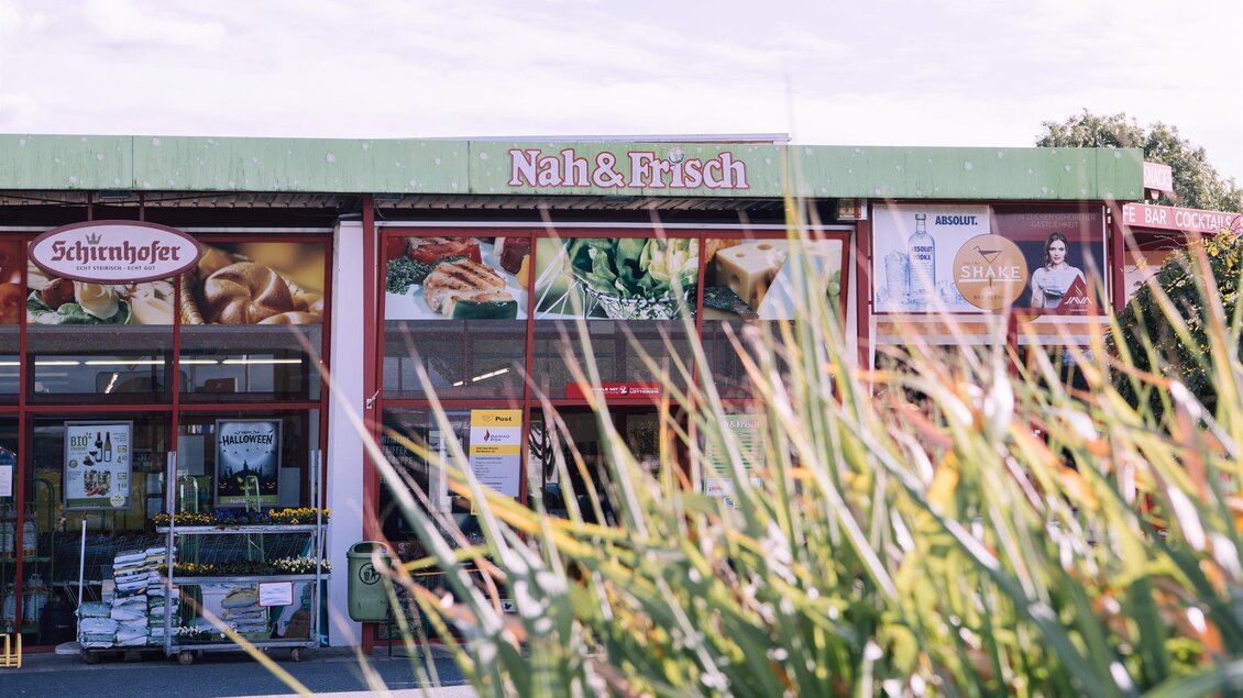 Ein Lebensmittelgeschäft mit dem Namen "Nah & Frisch". Die Fassade ist mit bunten Werbung und Pflanzen im Vordergrund dekoriert. | © Kurkommission Bad Blumau