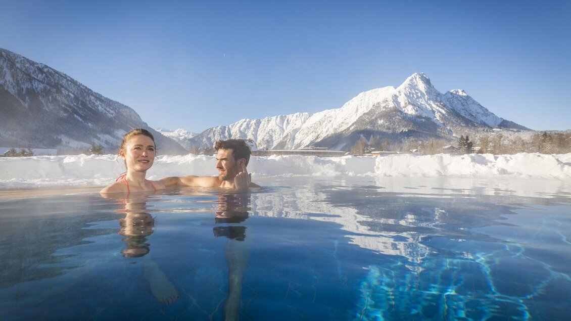 Ein entspannendes Thermalbecken in einer winterlichen Berglandschaft. Zwei Personen genießen die Aussicht auf schneebedeckte Berge unter klarem blue Himmel. | © Kujat
