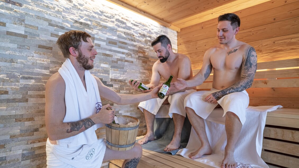 Drei Männer entspannen in einer Sauna. Sie genießen Getränke und haben Handtücher umgebunden.