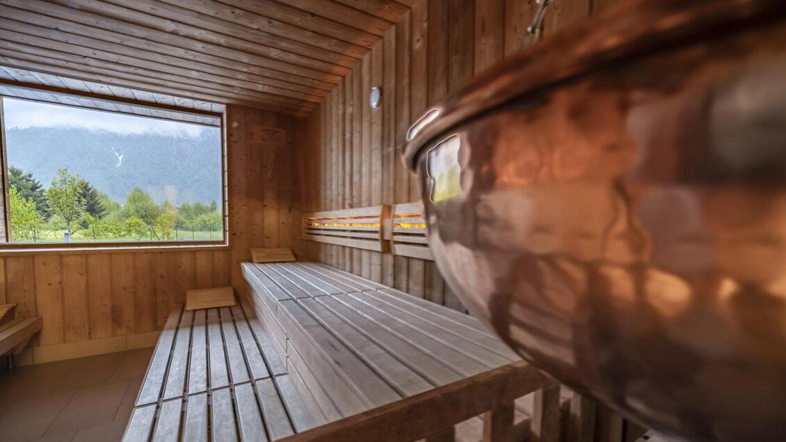 Eine moderne Sauna mit Holzwänden und einer großen Fensterfront, die einen Blick auf die Berge bietet. Im Vordergrund ist ein kupferner Kessel zu sehen. | © Thomas Lahnsteiner