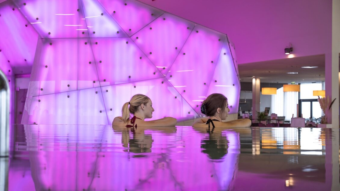 Zwei Frauen entspannen in einem modernen Pool mit schimmerndem Wasser. Im Hintergrund leuchtet eine große, geometrische Wand in sanften Pinktönen. | © Thomas Lahnsteiner