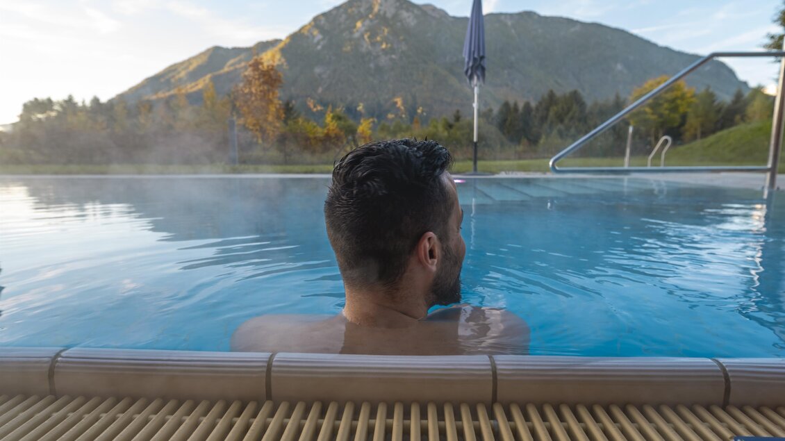 Ein entspannter Mann sitzt in einem warmen Pool mit Blick auf die Berge. Die Szenerie ist friedlich und einladend.