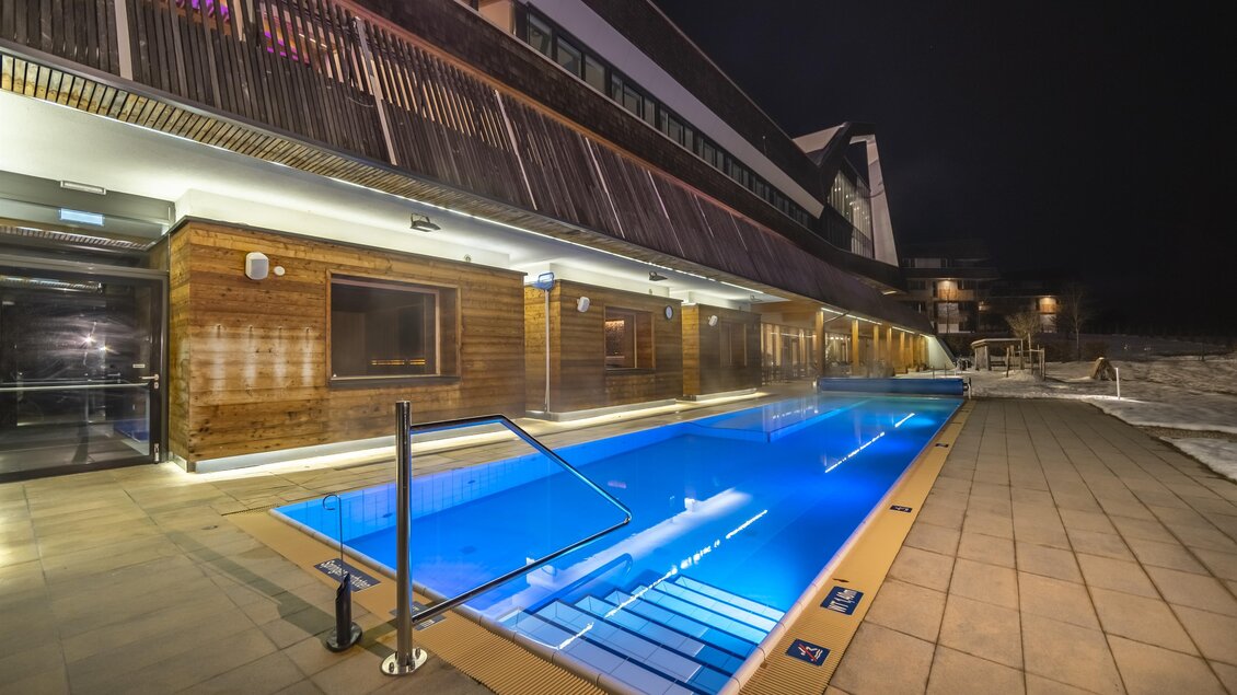 Ein eleganter Außenpool in der Nacht, umgeben von einer modernen Holzarchitektur. Das Wasser leuchtet blau und schafft eine entspannte Atmosphäre.