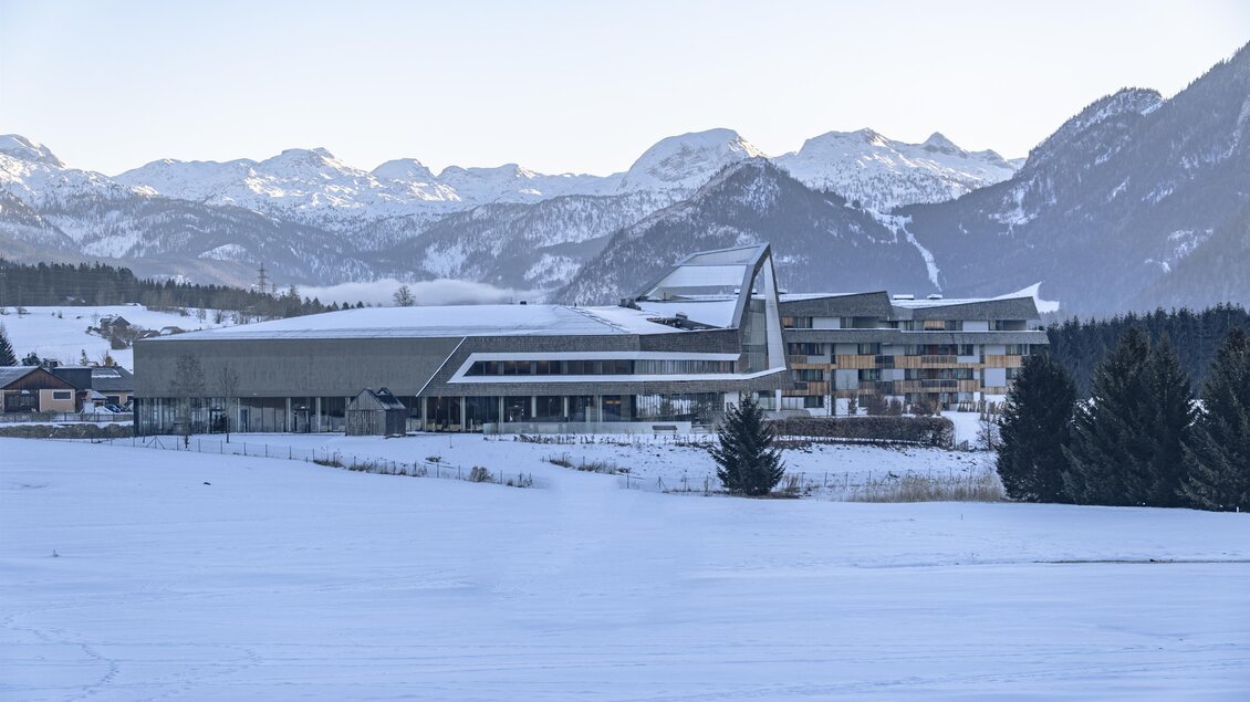 Ein modernes Gebäude liegt in einer schneebedeckten Landschaft mit majestätischen Bergen im Hintergrund. Die Szenerie strahlt Ruhe und winterliche Schönheit aus.