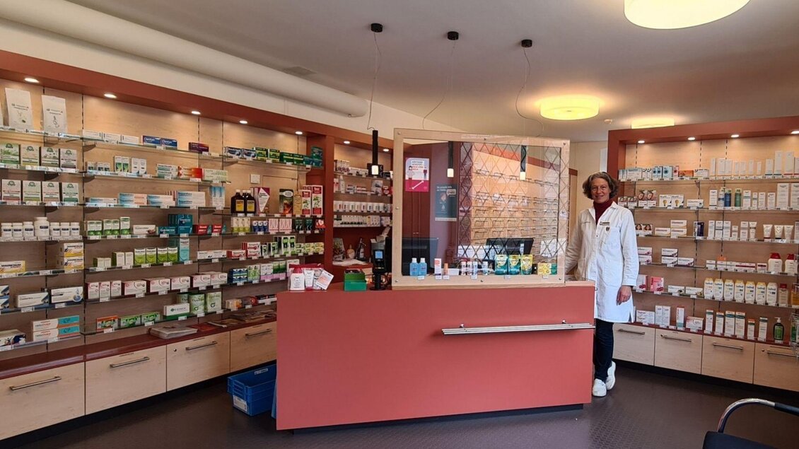 Ein freundlicher Apotheker steht an der Verkaufstheke in einer modernen Apotheke. Regale sind mit verschiedenen Medikamenten und Gesundheitsprodukten gefüllt. | © Stephanie Bor