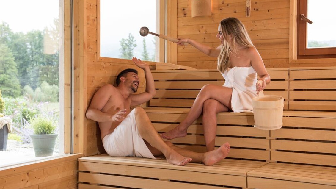 Ein Paar entspannt in einer Sauna. Die Frau hat einen Eimer und einen Schöpflöffel in der Hand. | © Hotel "Grüne Au"