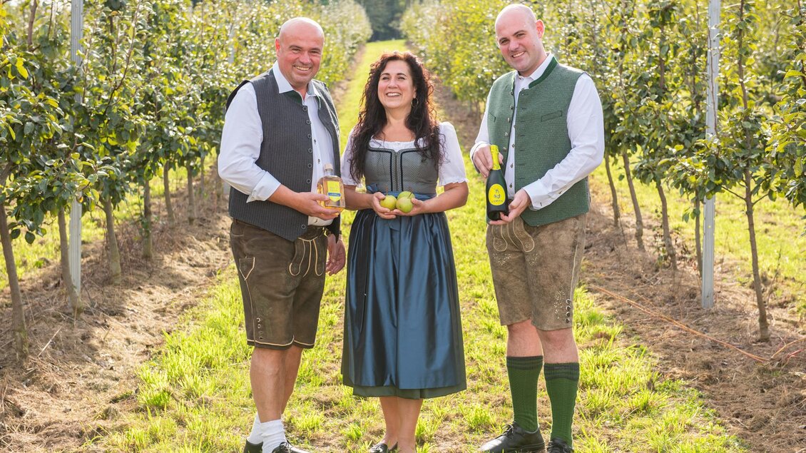 Drei Personen in traditioneller Kleidung stehen in einem Obstgarten. Sie halten Äpfel und Getränke in der Hand. | © Hotel "Grüne Au"