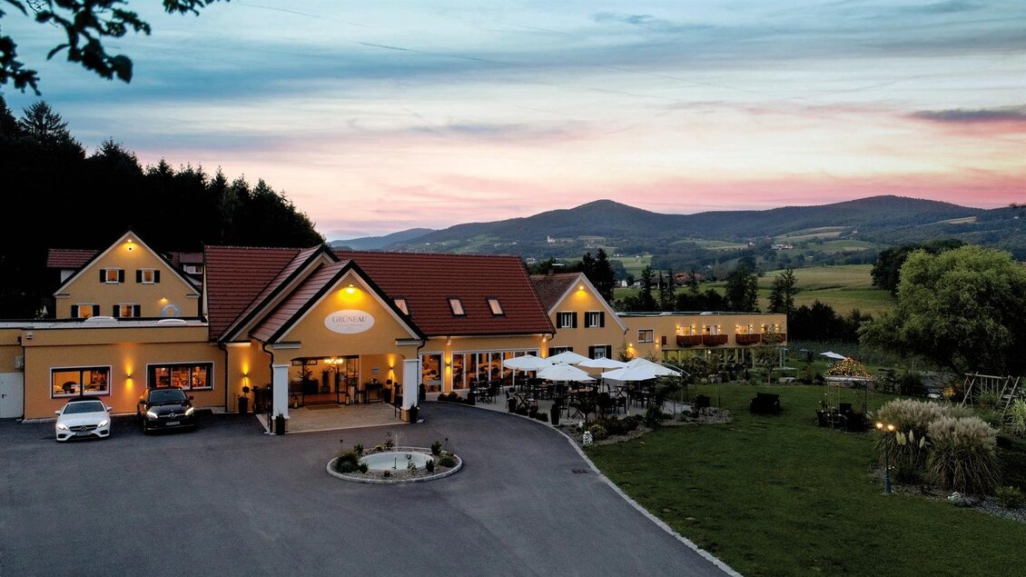 Ein charmantes Hotel mit rotem Dach befindet sich in einer malerischen Umgebung. Im Hintergrund sind sanfte Hügel und ein farbenfroher Himmel zu sehen. | © Hotel "Grüne Au"