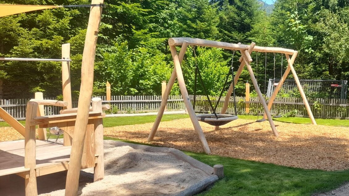 Ein schöner Spielplatz mit einer Holzschaukel und einer Spielstruktur. Der Bereich ist von Bäumen umgeben und hat eine grüne Wiese. | © Isabell Schachner