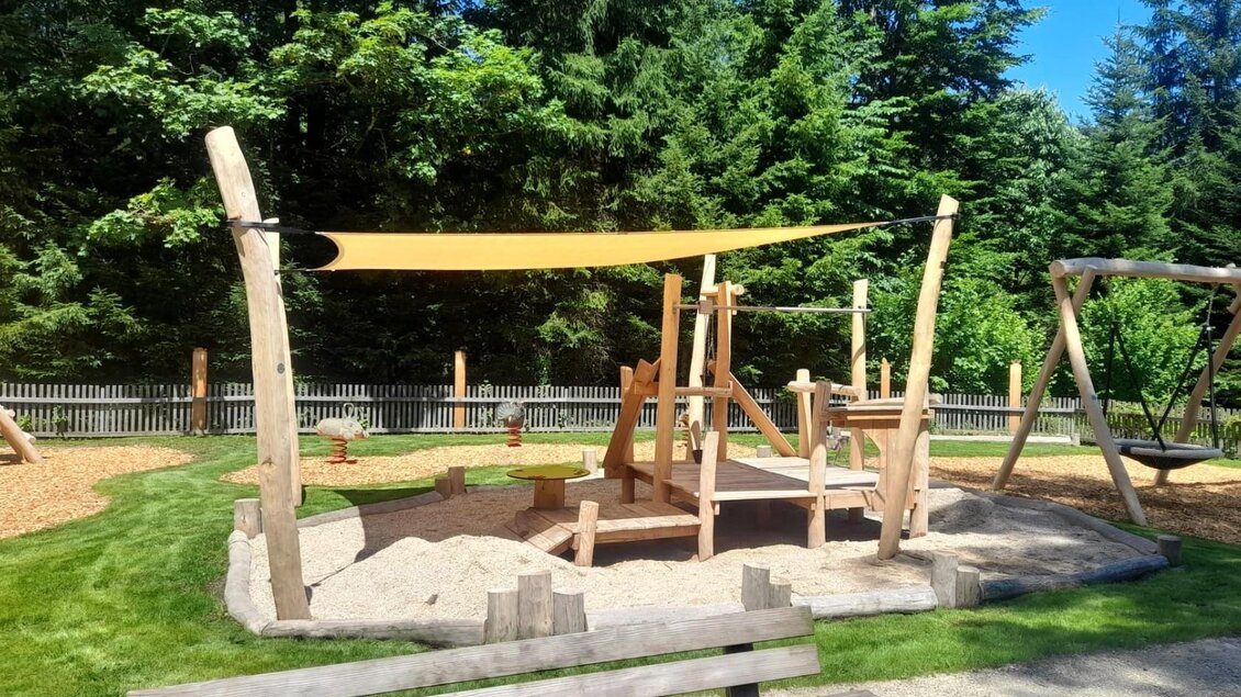 Ein moderner Spielplatz aus Holz mit einer Sandfläche und einem Sonnensegel. Im Hintergrund sieht man hohe Bäume und eine Schaukeln. | © Isabell Schachner