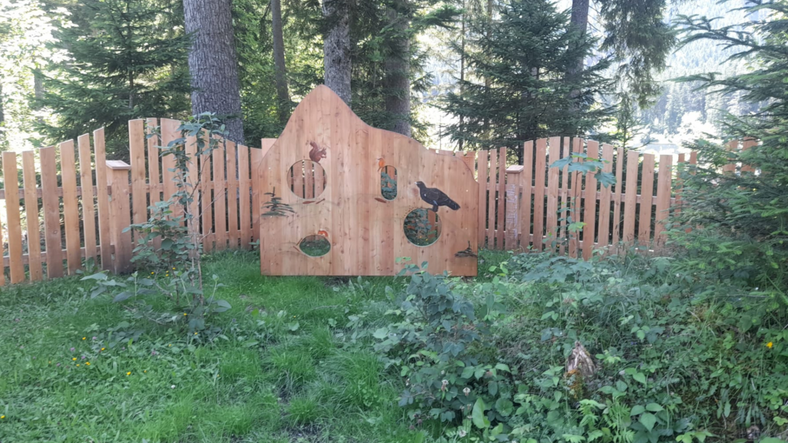 Eine hölzerne Wand mit Tiermotiven, umgeben von einer grünen Wiese. Im Hintergrund stehen hohe Bäume. | © Isabell Schachner