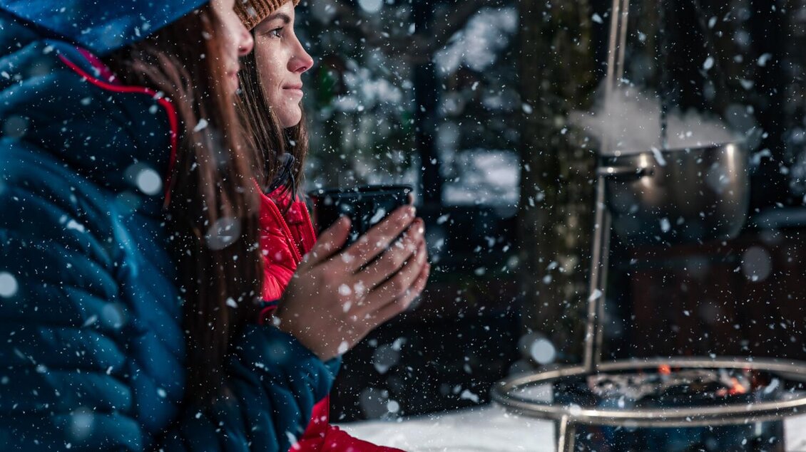 Zwei Personen in warmer Kleidung sitzen bei Schnee und trinken aus Tassen. Im Hintergrund ist ein Feuerstelle zu sehen, die für Wärme sorgt. | © Thomas Sattler