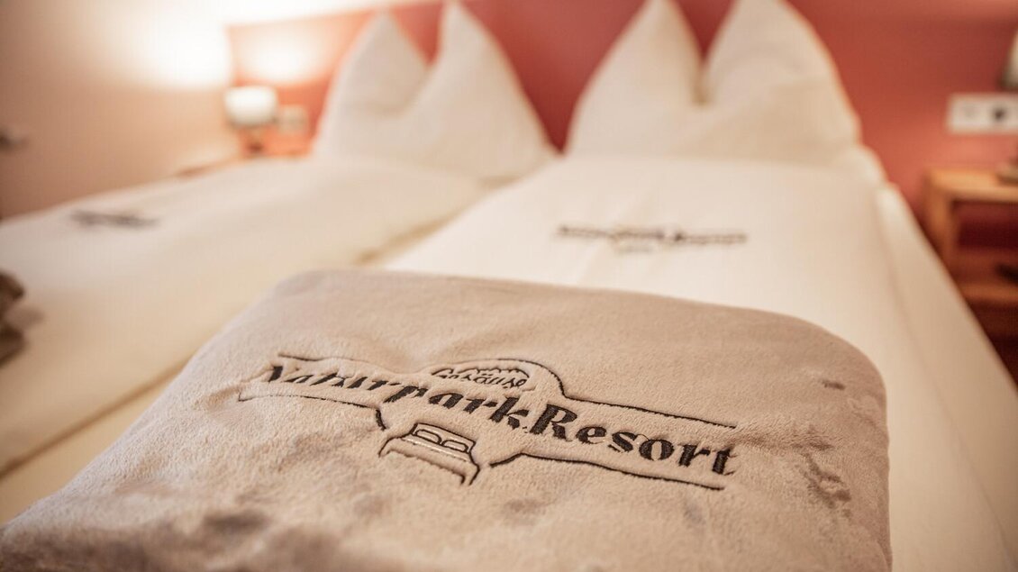 Ein gemütliches Hotelzimmer mit zwei Betten und weißen Kissen. Ein Handtuch mit dem Logo des Naturpark-Resorts liegt auf dem Bett. | © Thomas Sattler