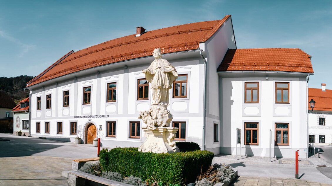 Ein historisches Gebäude mit einer roten Dachkonstruktion und großen Fenstern. Vor dem Gebäude steht eine Statue, umgeben von einer grünen Hecke. | © Stefan Leitner