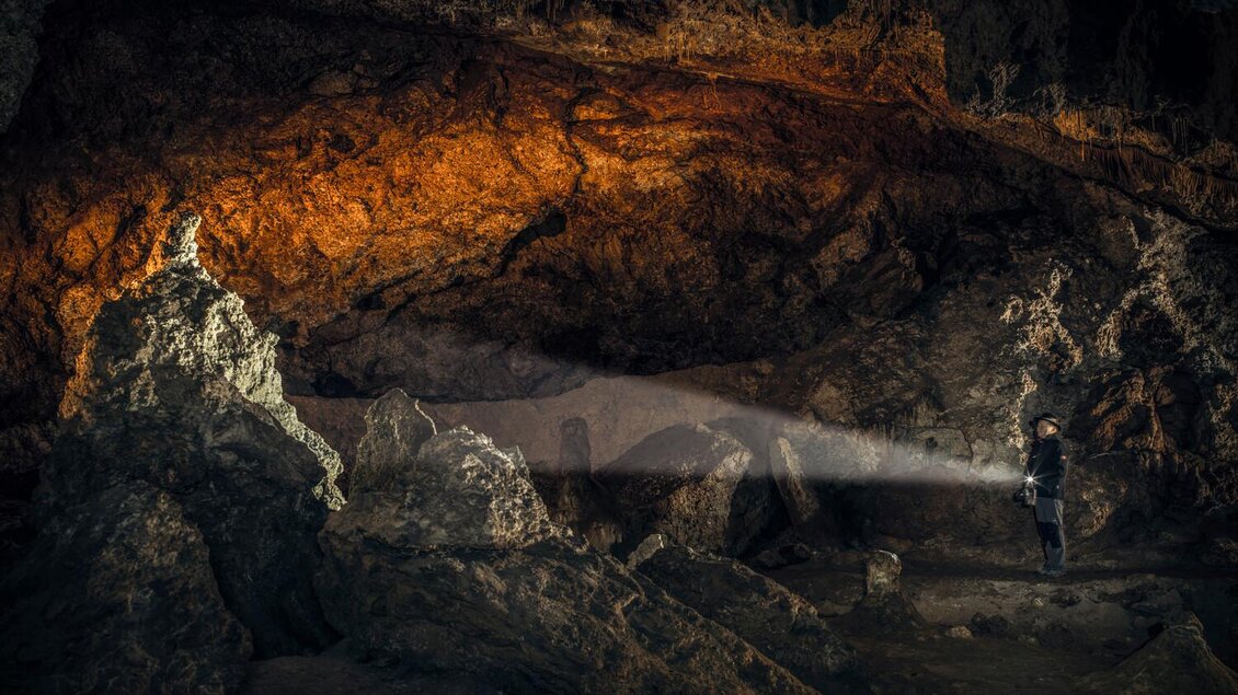 Ein Abenteurer steht in einer beeindruckenden Höhle und beleuchtet die Felsen mit einer Taschenlampe. Die Wände der Höhle schimmern in rot-orangefarbenen Tönen. | © Stefan Leitner