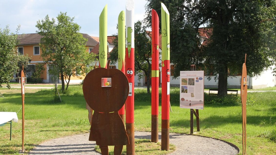 Eine künstlerische Installation mit bunten Stelen und einer metallenen Figur im Freien. Der Hintergrund zeigt Gebäude und Bäume in einer grünen Umgebung. | © OIKOS