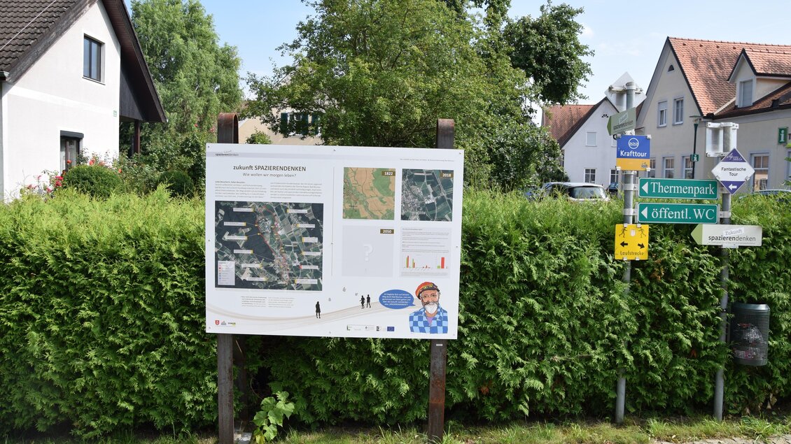 Ein Informationsschild steht in einer grünen Umgebung. Im Hintergrund sind Wohnhäuser und Straßenschilder sichtbar. | © Franz Spörk