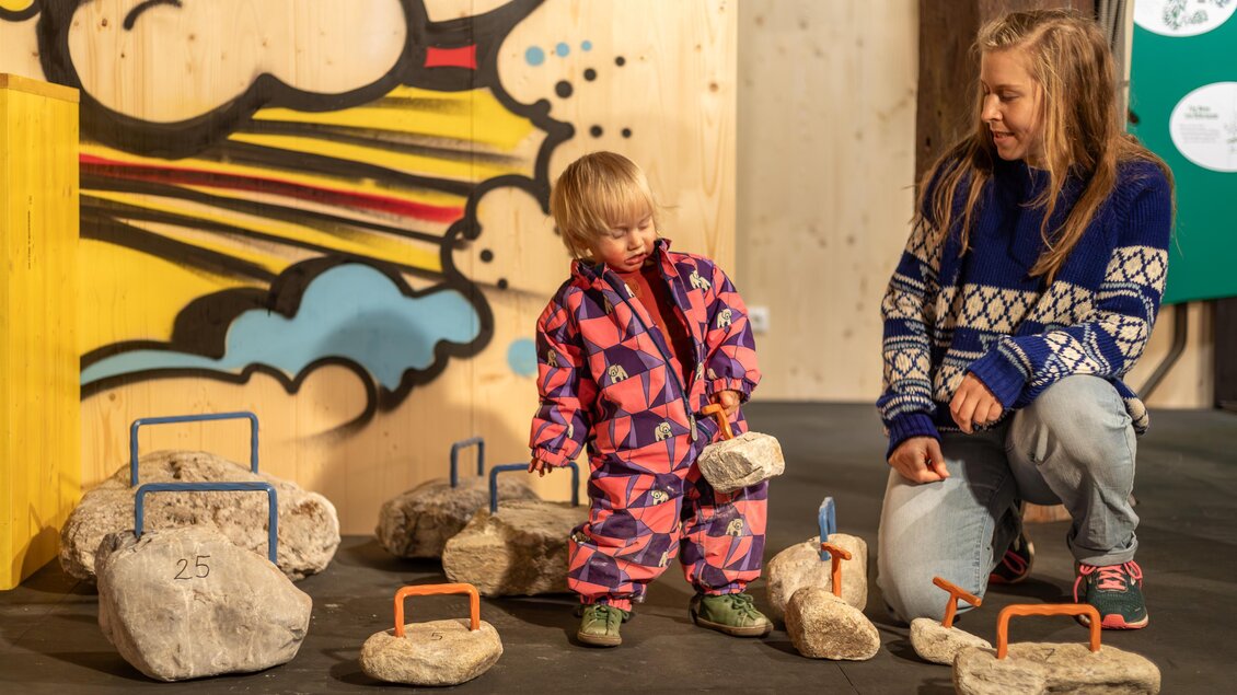 Ein kleines Kind in einem bunten Anzug spielt mit Steinen und hebt einen davon an. Neben ihm sitzt eine Frau und beobachtet ihn freundlich. | © Naturpark Mürzer Oberland