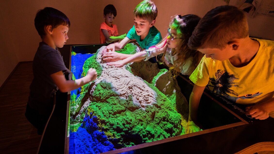 Eine Gruppe von Kindern gestaltet mit ihren Händen eine bunte Sandlandschaft in einem interaktiven Tisch. Die verschiedenen Farben leuchten und schaffen eine faszinierende Umgebung. | © Tourismusverband Murau