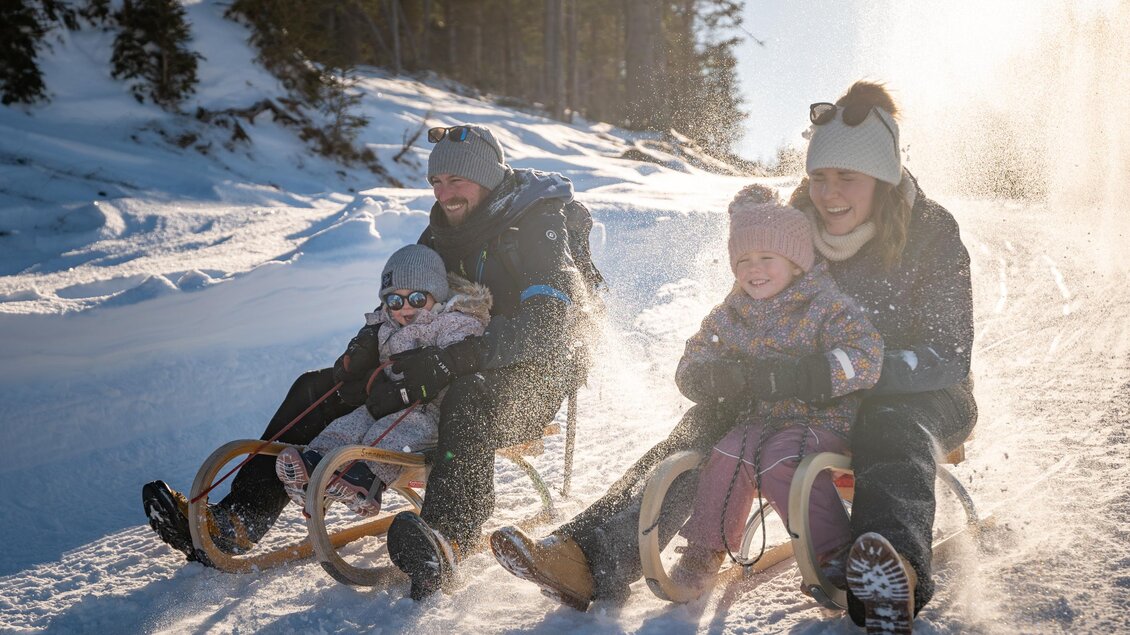 Eine Familie fährt mit Schlitten einen verschneiten Hang hinunter. Die Sonne scheint und es sieht nach viel Spaß aus. | © Oststeiermark Tourismus