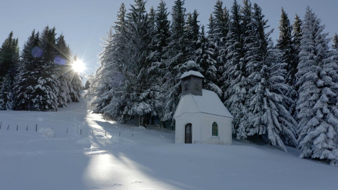 Eine kleine Kirche in einer verschneiten Landschaft, umgeben von hohen, schneebedeckten Bäumen. Die Sonne scheint während des Winters strahlend über die Szenarie. | © Foto Mellacher