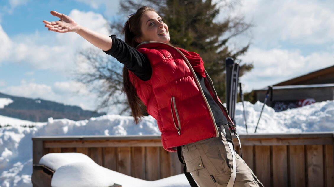Ein Mädchen steht fröhlich im Schnee und streckt die Arme aus. Im Hintergrund sind schneebedeckte Berge und ein Holzstapel zu sehen. | © Bernhard Bergmann