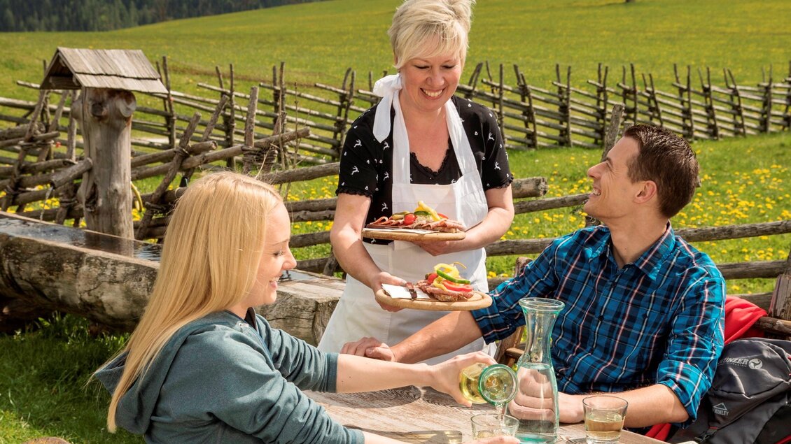 Eine freundliche Bedienung bringt Essen zu zwei Gästen im Freien. Die Umgebung ist ländlich mit einer schönen Wiese und einem bewaldeten Hintergrund. | © Naturpark Almenland