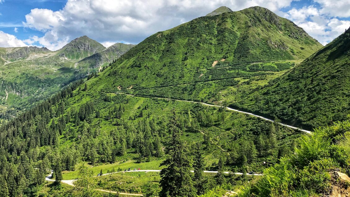Eine grüne Berglandschaft mit sanften Hügeln und bewaldeten Tälern. Der Himmel ist blau mit weißen Wolken.