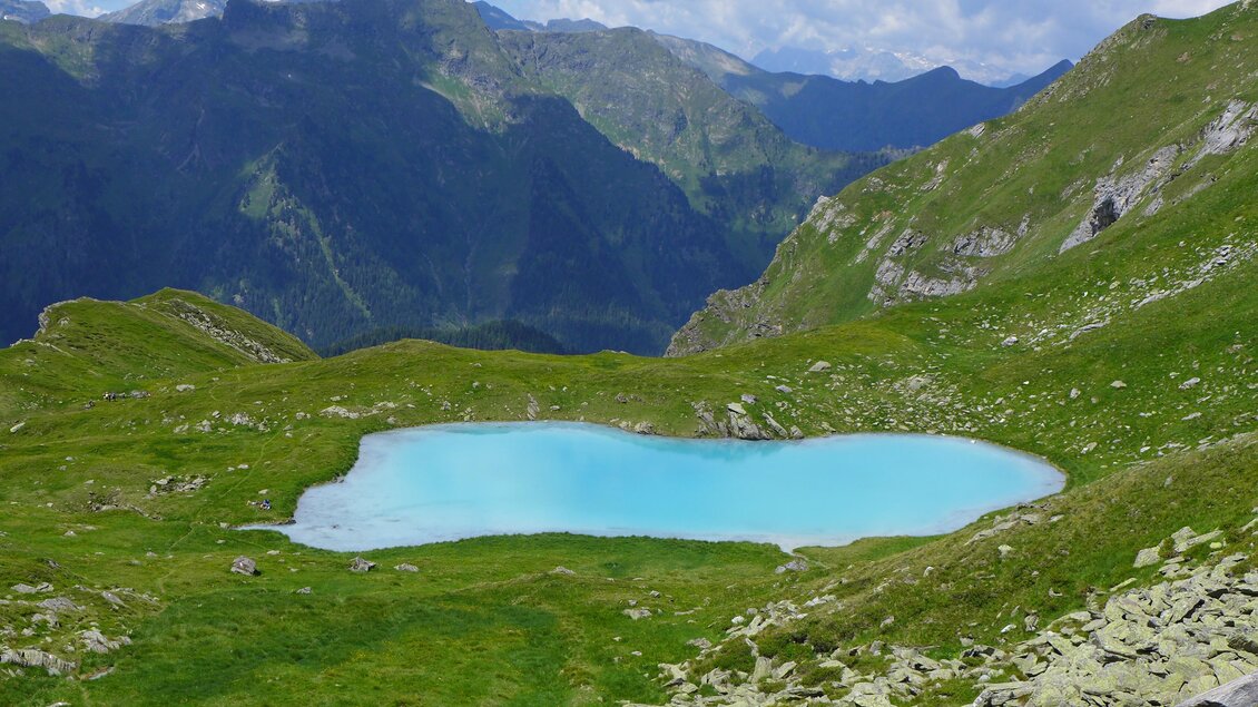 Ein idyllischer Bergsee mit klarem, türkisfarbenem Wasser, umgeben von grünen Wiesen und Bergen. Der Himmel ist teilweise bewölkt, was eine friedliche Atmosphäre schafft.