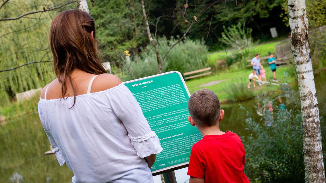 Eine Frau und ein Kind lesen ein Informationsschild am Teich. Im Hintergrund sind weitere Personen, die in der Natur aktiv sind. | © Gemeinde Unterlamm