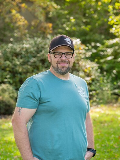 A man is standing outside in a green park. He is wearing a blue T-shirt and a black cap, with trees in the background. | © Helmut Schweighofer