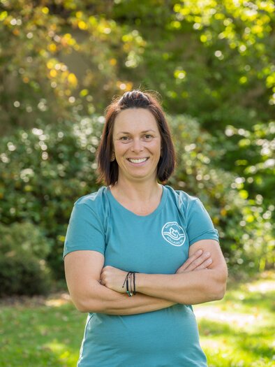 A smiling woman with short dark hair is standing in a green park. She is wearing a blue T-shirt and has her arms crossed. | © Helmut Schweighofer