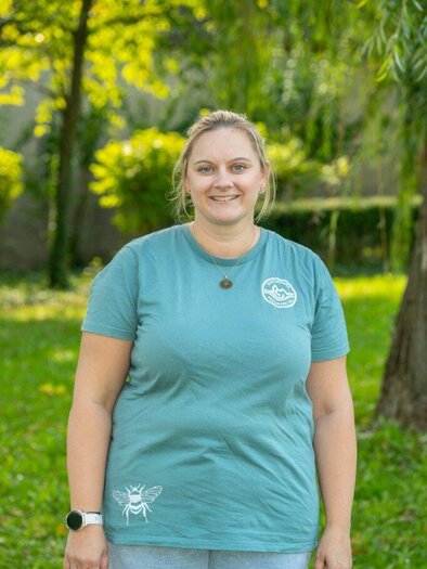 A smiling woman sits in front of a background with a green meadow. She is wearing a green T-shirt and appears friendly. | © Helmut Schweighofer