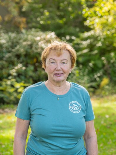 An older woman is standing in a meadow with trees in the background. She is wearing a blue t-shirt and is smiling warmly. | © Helmut Schweighofer