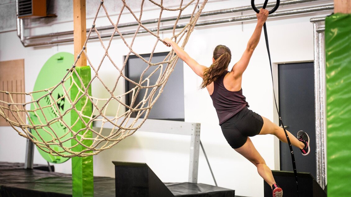Frau klettert an einem Ninja Warrior Parcours in der Ninjabox Graz, während sie sich an einem Seil festhält und über ein Netz schwingt. | © Ninjabox_7