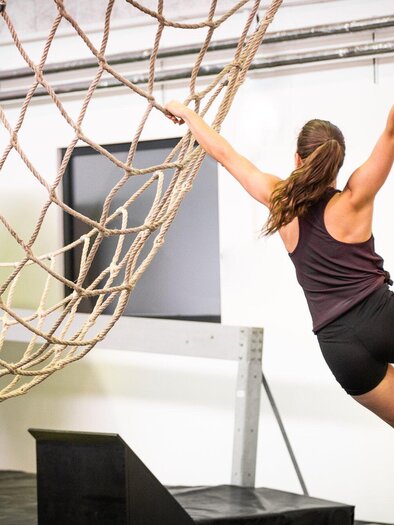 A woman climbs on a Ninja Warrior course in the Ninjabox Graz, while holding onto a rope and swinging over a net. | © Ninjabox_7
