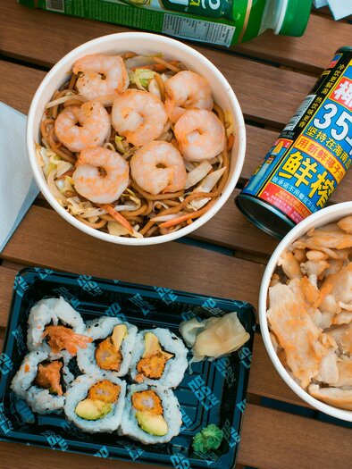 A tray with various Asian dishes, including noodles with shrimp, fried chicken, and sushi. Accompanied by a drink in a colorful can. | © Noodle King – Michael Choup
