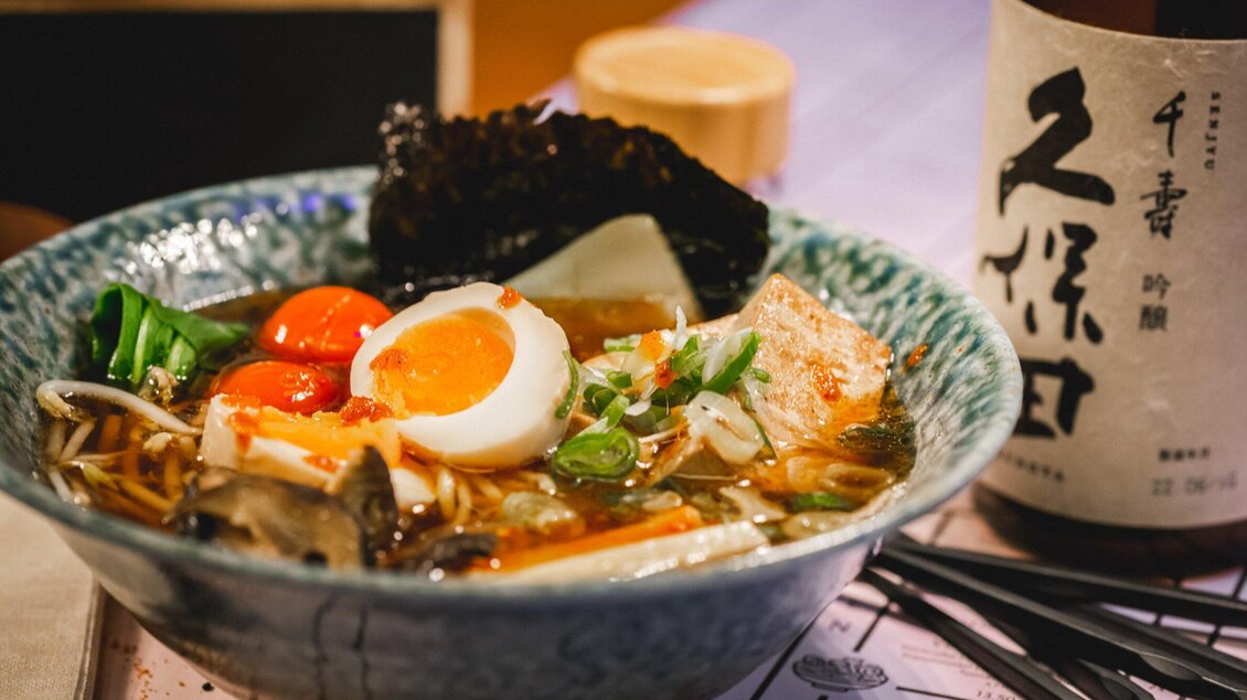 Eine Schüssel Ramen mit weichen Eiern, Gemüse und Algen. Neben der Schüssel steht eine Flasche Sake. | © Noonbar - Moriwano Group