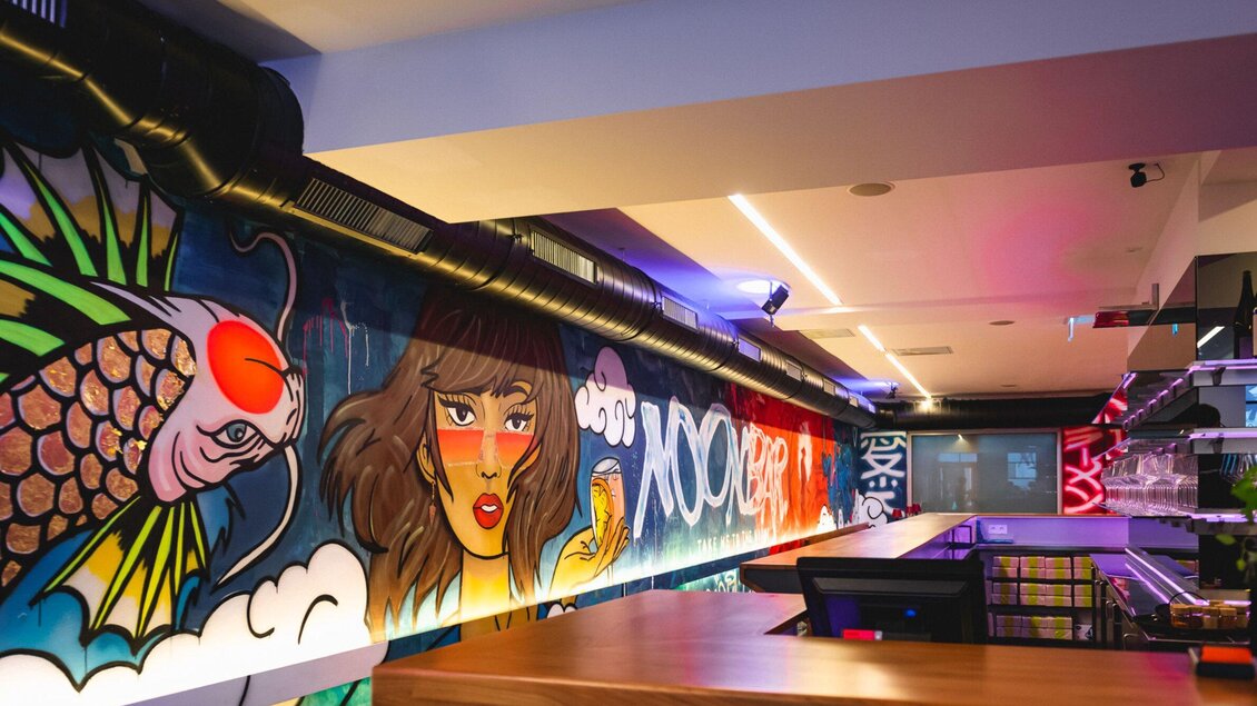 Ein moderner Innenraum mit einer auffälligen Wandgestaltung aus buntem Graffiti. Die Bilder zeigen stilisierte Fische und eine Frau in lebhaften Farben. | © Noonbar - Moriwano Group
