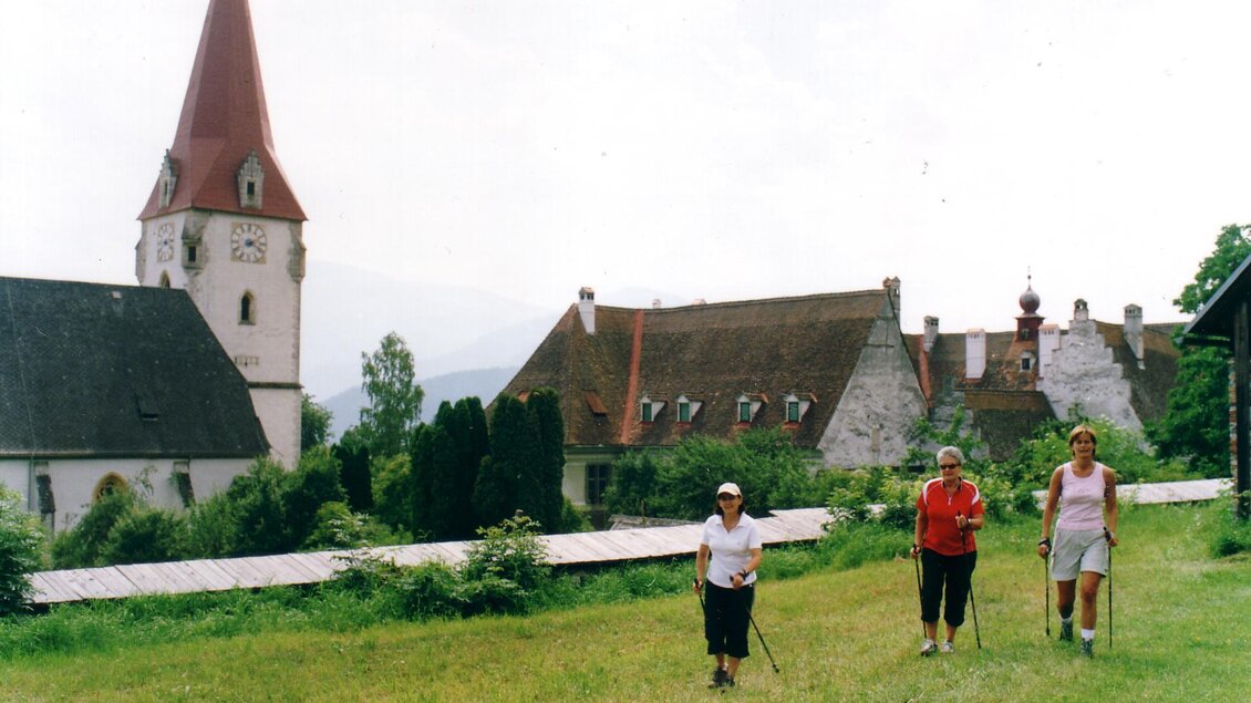 Drei Personen wandern auf einer Wiese in der Nähe eines historischen Gebäudes mit einem hohen Kirchturm. Die Landschaft ist grün und zeigt eine ruhige, ländliche Umgebung.