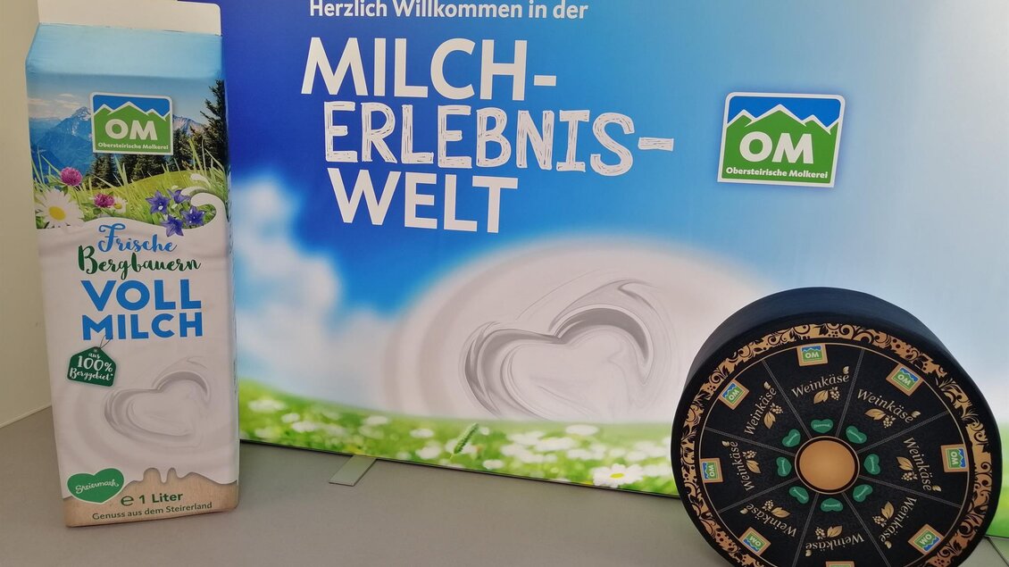 Ein Willkommensschild für die Milch-Erlebniswelt mit dem Logo von OM. Daneben steht eine Verpackung mit frischer Vollmilch und eine runde Tafel. | © Obersteirische Molkerei
