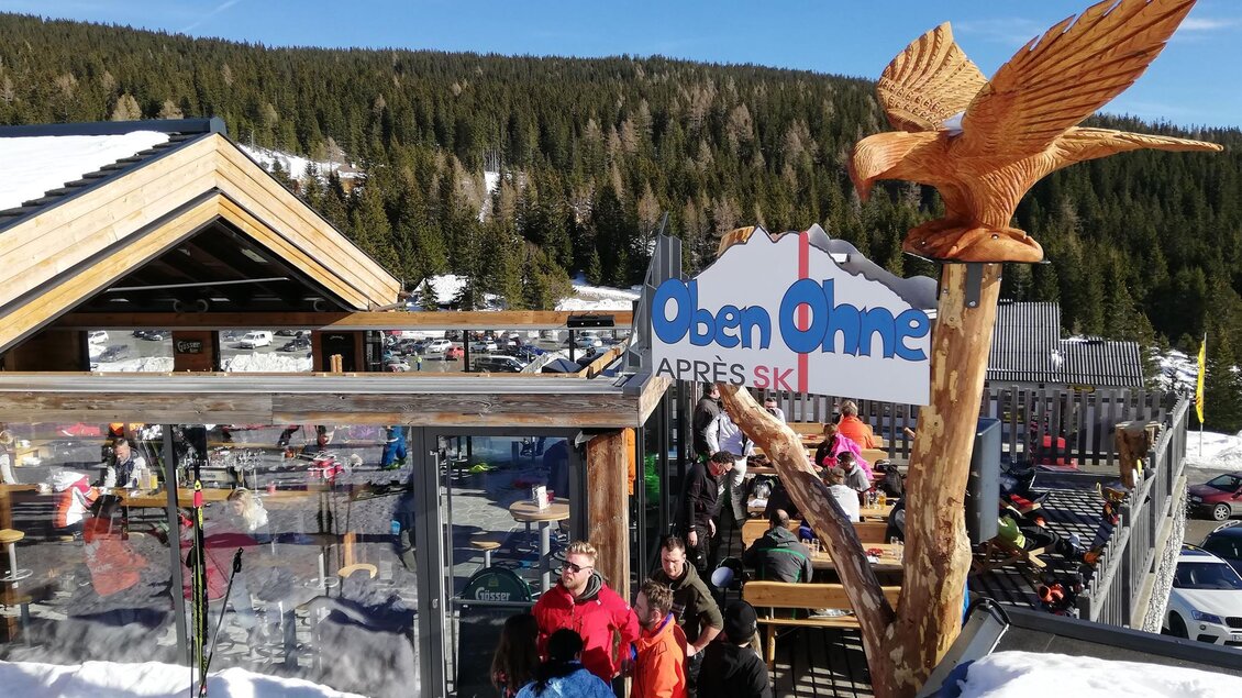 Ein lebendiges Après-Ski-Lokal mit Snowboardern und Skifahrern im Freien. Im Hintergrund erstrecken sich schneebedeckte Berge unter blauem Himmel. | © Oben Ohne - Aprés Ski