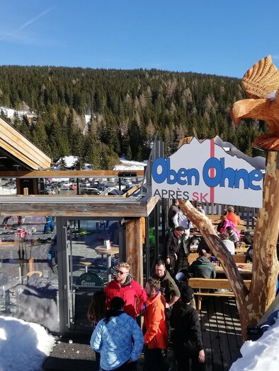 A lively après-ski venue with snowboarders and skiers outdoors. In the background, snow-covered mountains stretch under a blue sky. | © Oben Ohne - Aprés Ski