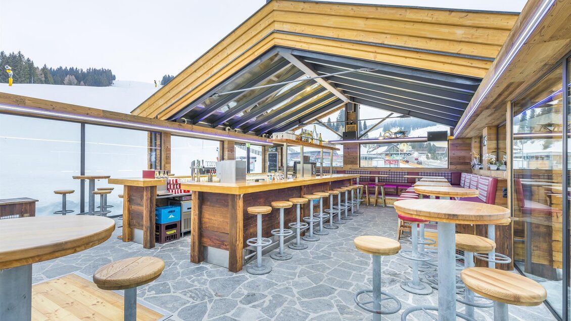 Eine moderne Bar mit Holzmöbeln und großen Glasflächen. Der Blick reicht über die Berge und die umliegende Landschaft. | © Oben Ohne - Aprés Ski