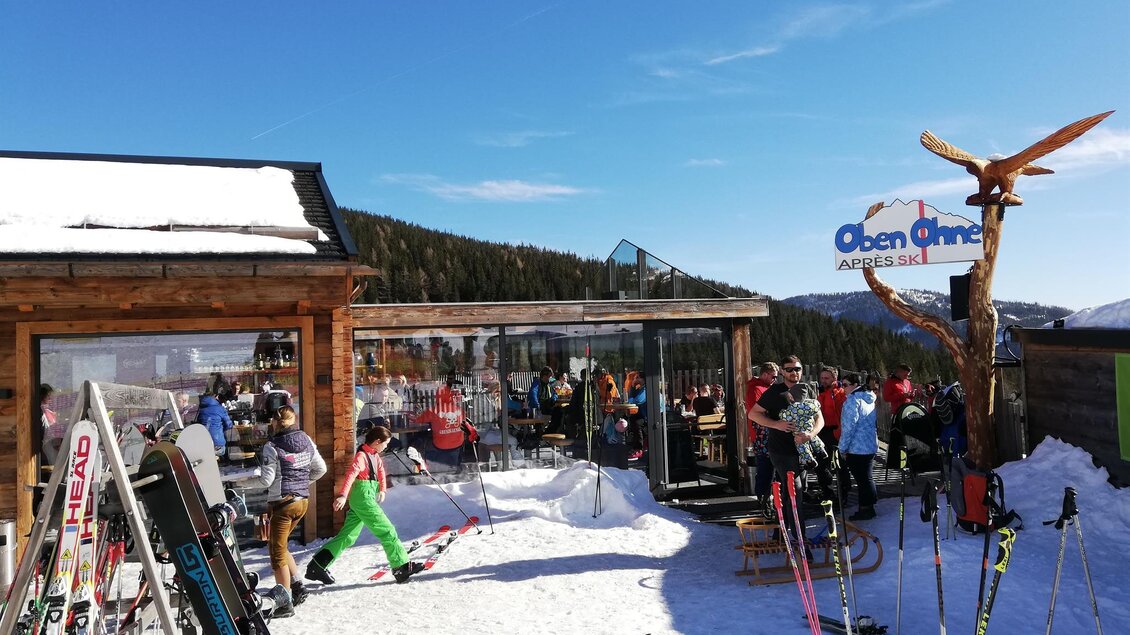 Eine Skihütte in den Bergen, umgeben von Schnee. Skifahrer und Besucher genießen die Sonne und entspannen im Freien. | © Oben Ohne - Aprés Ski