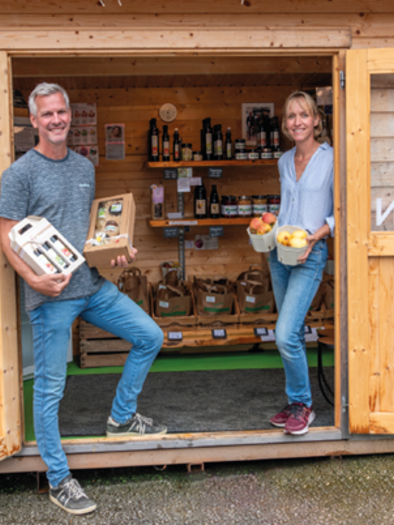 Eine Holzlade mit zwei Personen, die regionale Produkte präsentieren. Im Hintergrund sind Regale mit verschiedenen Lebensmitteln zu sehen. | © Mauthner