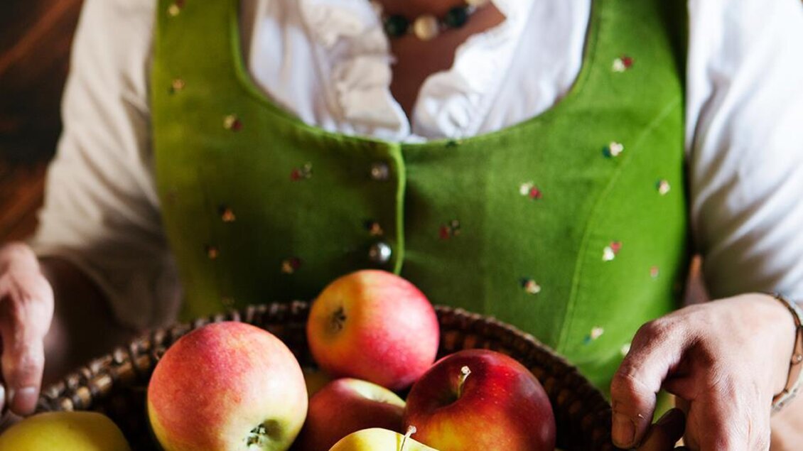 Eine Frau in traditioneller Tracht hält einen Korb mit verschiedenen Äpfeln. Die Äpfel sind bunt und frisch, und sie lächelt freundlich. | © Tourismusverband Thermen-Vulkanland