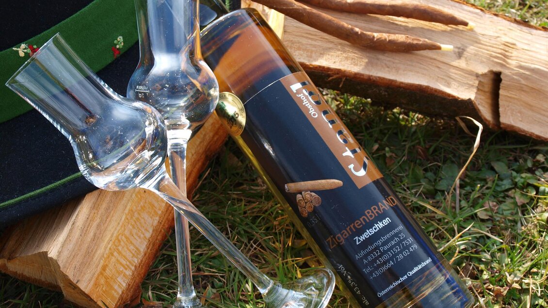Eine Flasche Schnaps steht neben zwei Gläsern auf einem Holzhaufen. Im Hintergrund ist ein Hut mit grüner Borte zu sehen. | © Obsthof Stangl