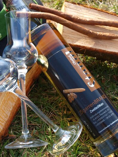 Eine Flasche Schnaps steht neben zwei Gläsern auf einem Holzhaufen. Im Hintergrund ist ein Hut mit grüner Borte zu sehen. | © Obsthof Stangl